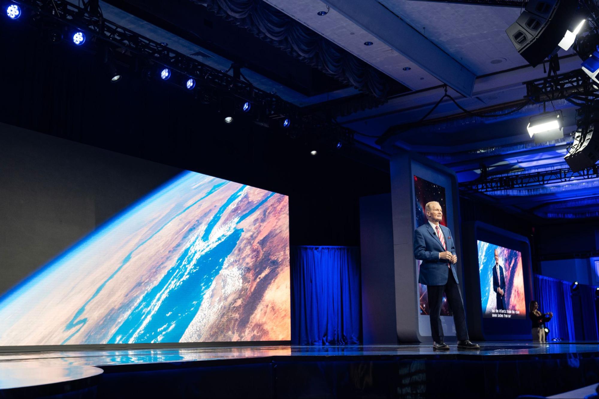 A presenter in a suit stands on a dimly lit stage in front of a large screen showing Earth from space.