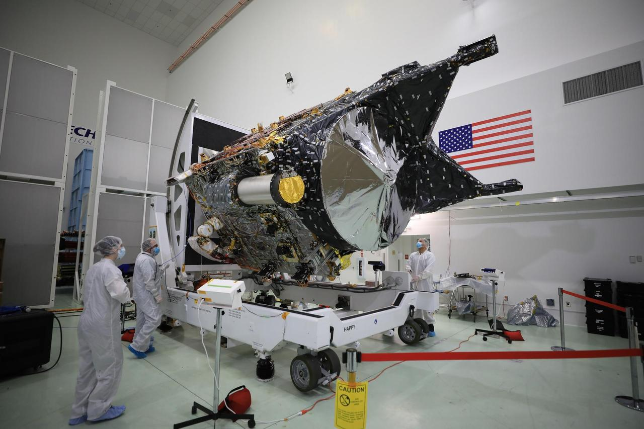 Large spacecraft covered in foil in clean room with technicians and American flag.