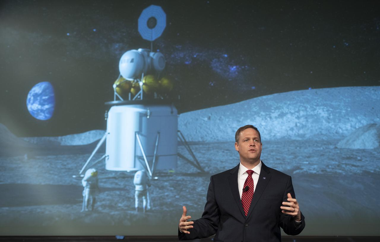 Man in suit gestures before screen displaying lunar lander and Earth.
