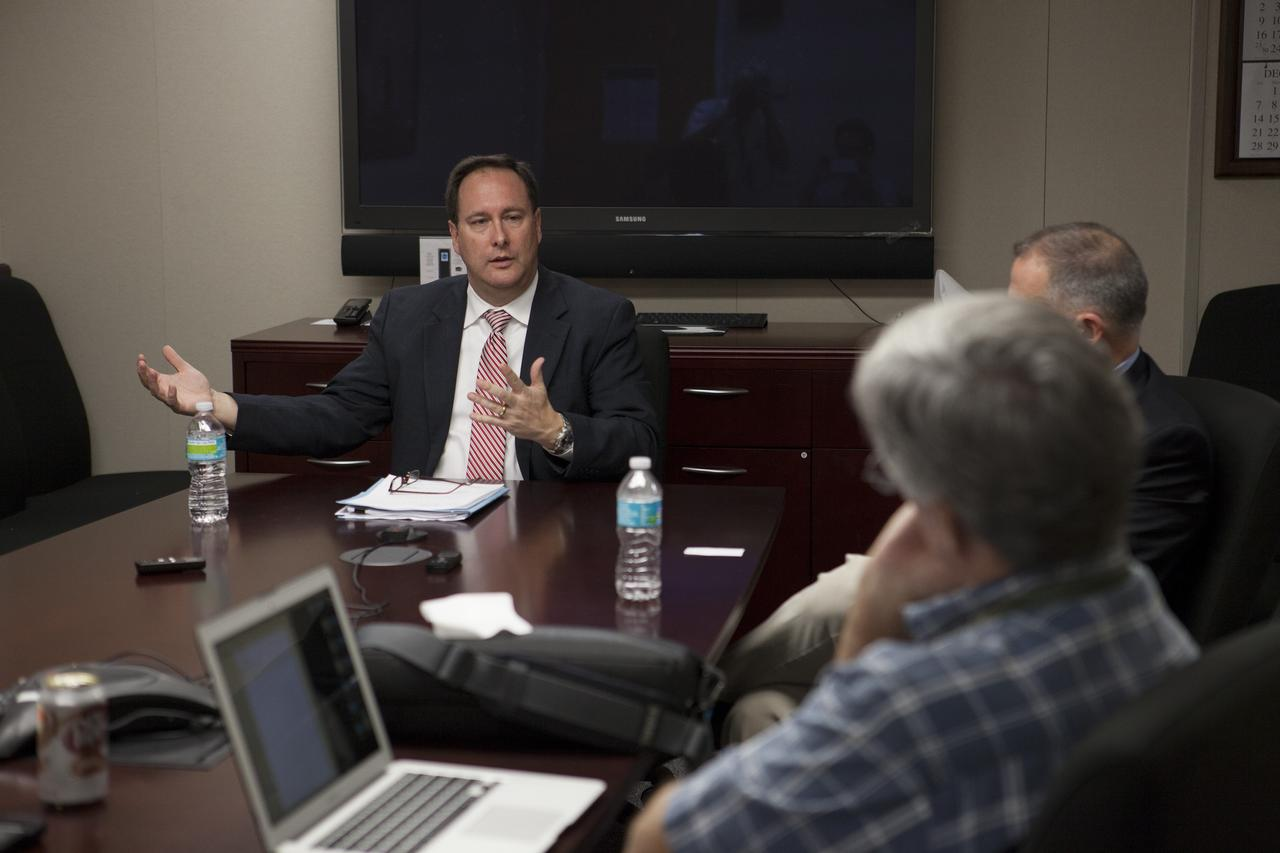 Man in suit speaking and gesturing at conference table with others listening.