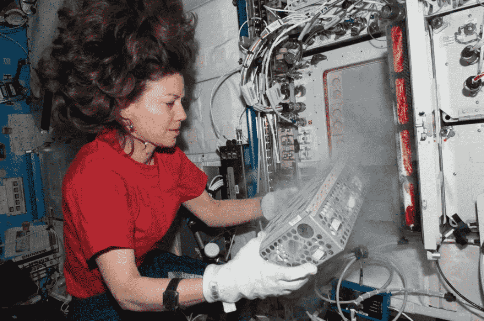 Woman in microgravity with floating hair handles equipment wearing gloves amid wires and panels
