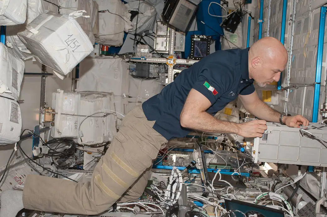 Man in blue shirt and beige pants works in cluttered space station module with cables